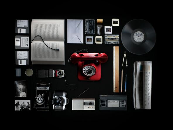 Anordnung verschiedener Medien auf einem schwarzen Hintergrund. Zu sehen sind ein altes rotes Telefon, Schallplatten, ein Buch, eine Kamera, eine Videokassetten, Disketten, ein kleines tragbares Radio sowie Fotos und weitere Medien.