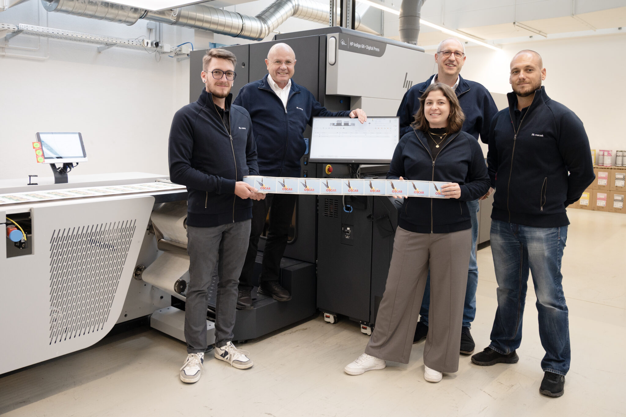Dr. Johannes Michael Wareka (CEO, 4. Generation der Gründerfamilie), Stefanie Wareka (CMO und 5. Generation der Gründerfamilie), COO Michael Grech und Mitarbeiter der Produktion Digital Offset mit der HP Indigo 6k+ Digital-Offsetmaschine, der jüngsten Innovation im Maschinenpark von Marzek Etiketten+Packaging.